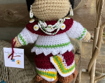 Puede incluir: Muñeca de ganchillo hecha a mano con cabello negro, vestida con un atuendo blanco y granate con detalles en verde y amarillo. La muñeca tiene joyas de cuentas y una etiqueta que dice "Sandy's Country Crochet". La muñeca se exhibe sobre una superficie de madera.