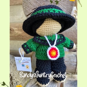 May include: Crocheted doll wearing a green sweater, black trousers, and a black hat with a green band. The doll is holding a white tag with the text "Sandy's Country Crochet" and a small image of three birdhouses.
