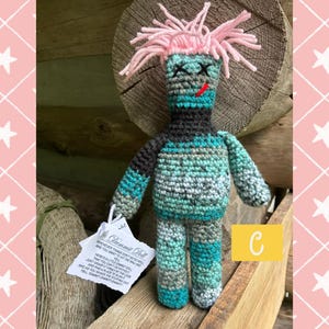 May include: A crocheted doll with pink yarn hair, black stitched X eyes, and a red stitched mouth. The body features a mix of teal, grey, and black yarn. A white tag attached to the arm reads Dammit Doll with text explaining its use.