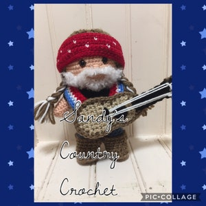 May include: A crocheted doll wearing a red bandana, a white beard, and holding a guitar. The doll is wearing blue jeans and brown boots. The text "Sandy's Country Crochet" is written below the doll.