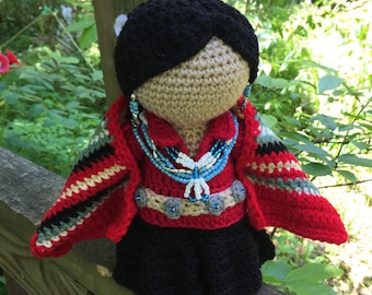 May include: Crocheted doll with black hair, wearing a red dress with a beaded necklace and a colorful shawl. The doll is standing on a wooden surface.
