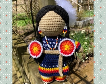 May include: Crocheted doll wearing a blue dress with red, yellow, and orange accents. The doll has black hair, a white feather, and beaded earrings. The doll is holding two red, yellow, and orange crocheted circles.