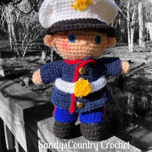 May include: A crocheted doll dressed as a US Marine. The doll is wearing a blue uniform with a white belt and a yellow emblem. The doll is wearing a white hat with a black brim and a yellow emblem.