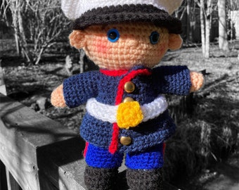 May include: A crocheted doll dressed as a US Marine. The doll is wearing a blue uniform with a white belt and a yellow emblem. The doll is wearing a white hat with a black brim and a yellow emblem.