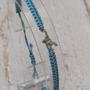 May include: A light blue macrame bracelet with a silver-colored monkey charm. The bracelet is displayed on a clear stand, showcasing the woven cord design and adjustable closure. The monkey charm hangs from the bracelet.