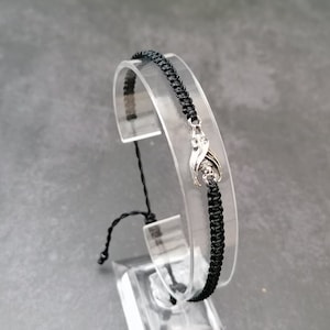May include: A black braided bracelet with a silver charm featuring two intertwined figures.