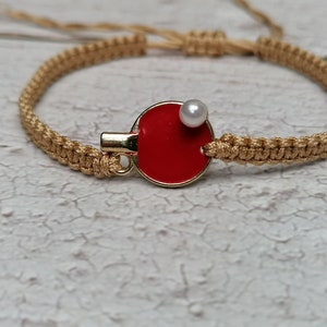 May include: A macrame bracelet with a gold-colored clasp and a red ping pong paddle charm. A small white pearl is attached to the paddle. The bracelet is made of tan-colored woven cord.