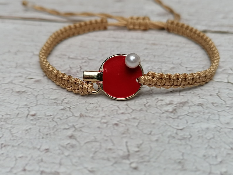 May include: A macrame bracelet with a gold-colored clasp and a red ping pong paddle charm. A small white pearl is attached to the paddle. The bracelet is made of tan-colored woven cord.