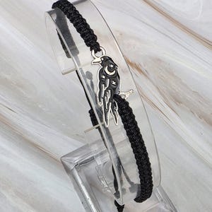 May include: A black woven cord bracelet featuring a silver-toned metal charm of a raven. The raven charm is decorated with black enamel details, a small crescent moon, and tiny star shapes on its body, attached to the center of the adjustable cord.