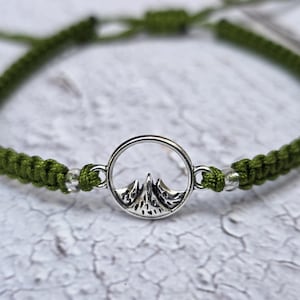Könnte beinhalten: Olivgrünes Makramee-Armband mit einem silberfarbenen Bergketten-Anhänger. Das Armband hat einen verstellbaren Schiebeknotenverschluss. Der Anhänger zeigt eine detaillierte Bergkette in einem kreisförmigen Rahmen.