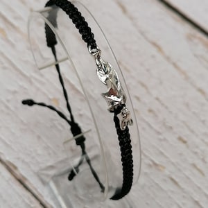Op de afbeelding: Een zwarte koordarmband met een zilverkleurige metalen vleermuisbedel. De armband is gemaakt met een gevlochten koordontwerp en bevat verstelbare schuifknopen aan het uiteinde om de pasvorm rond de pols vast te zetten.