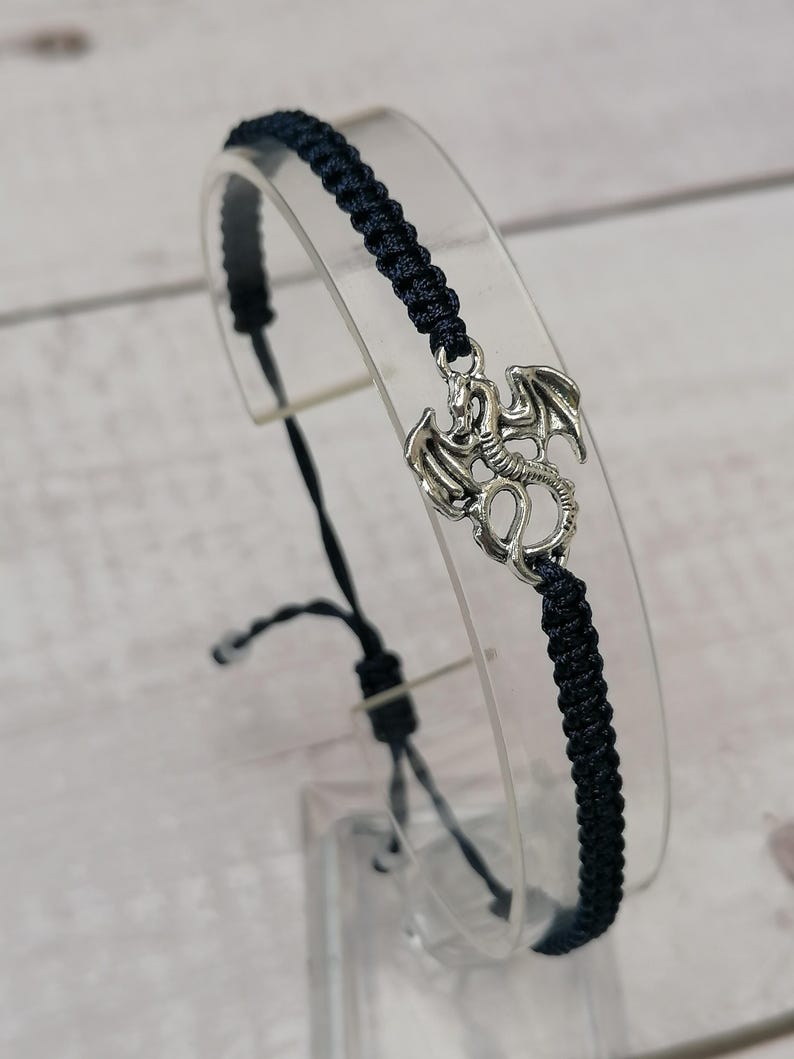 May include: A black braided bracelet with a silver dragon charm. The adjustable bracelet is shown on a clear stand against a light wood-grain background. The dragon charm has detailed wings and a serpentine body.