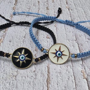 May include: Two braided cord bracelets with circular charms. One bracelet is black, the other is light blue. Each charm features a starburst design with an evil eye symbol in the center, set against a black or white background.
