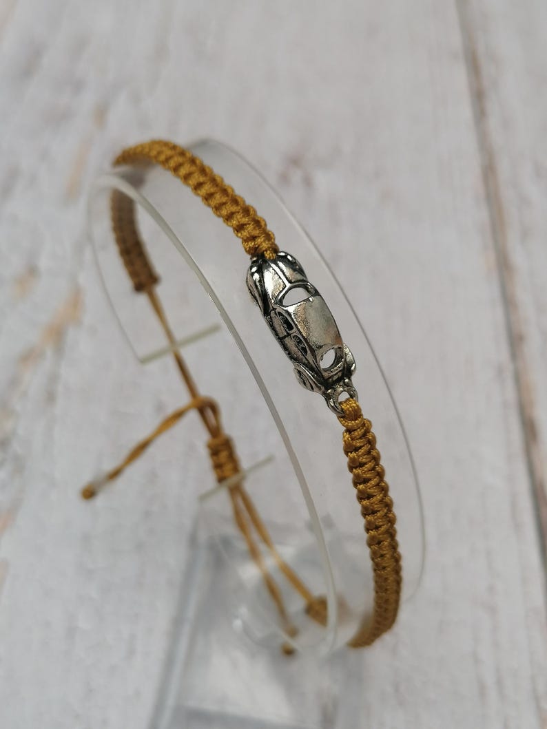 Puede incluir: Pulsera de macram&eacute; color mostaza con un dije de coche plateado. La pulsera es ajustable con nudos corredizos. El dije del coche est&aacute; detallado con ventanas y cap&oacute;. La pulsera se exhibe sobre un soporte transparente y curvado.