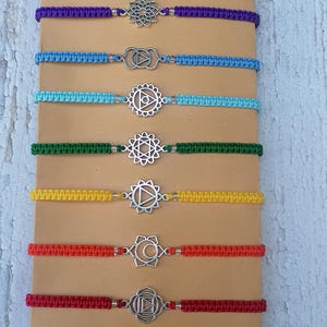 Op de afbeelding: Zeven chakra-armbanden met zilveren bedels op een bruine achtergrond. De armbanden zijn in de kleuren van de chakra's: paars, blauw, lichtblauw, groen, geel, oranje en rood.
