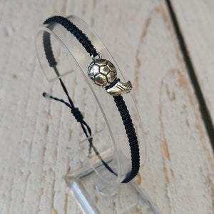 Könnte beinhalten: Schwarzes geflochtenes Armband mit einem silbernen Fussball-Anhänger.