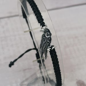 Puede incluir: Pulsera trenzada negra con un dije de plata de un cuervo con una luna creciente y estrellas en sus alas.