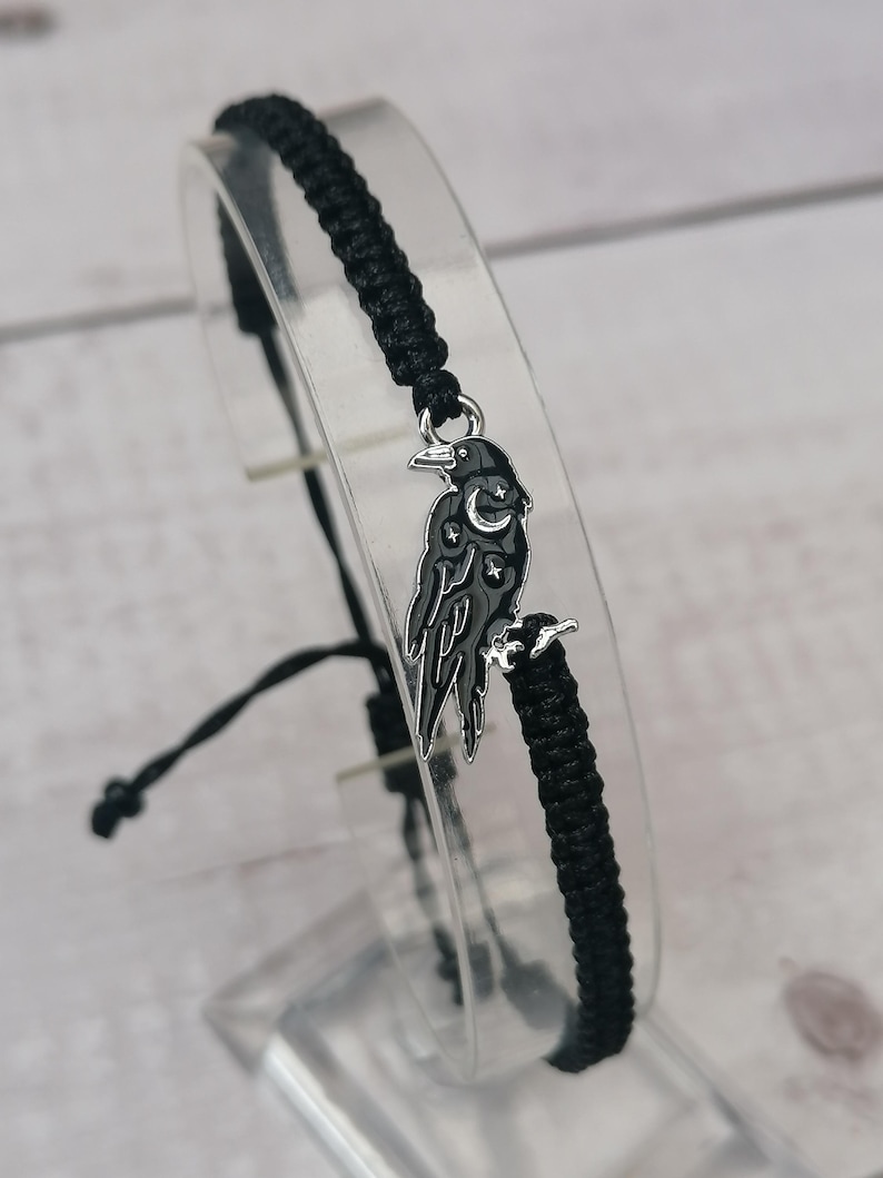 May include: Black braided bracelet with a silver charm of a raven with a crescent moon and stars on its wings.