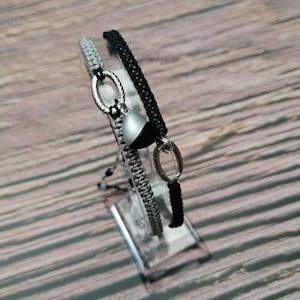 May include: Two braided bracelets with silver metal accents and a silver charm. One bracelet is black and the other is grey.