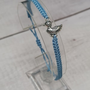 Könnte beinhalten: Ein hellblaues geflochtenes Armband mit einem silbernen Entenanhänger.