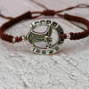 May include: A brown braided bracelet with a silver charm featuring a stylized axe design.