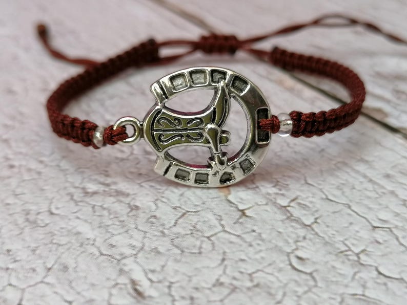 May include: A brown braided bracelet with a silver charm featuring a stylized axe design.