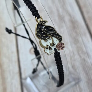 May include: Black braided bracelet with a raven and skull pendant. The raven is perched on a skull with a rose. The pendant has gold accents. The bracelet is displayed on a clear stand against a light wood background.