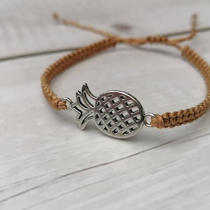 Puede incluir: Un brazalete de cordón trenzado marrón con un dije de piña plateado.