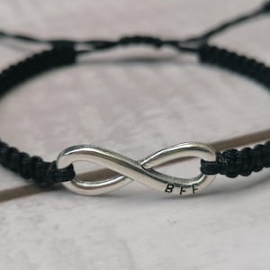 Puede incluir: Pulsera de cordón trenzado negro con un dije de infinito plateado que dice "BFF".