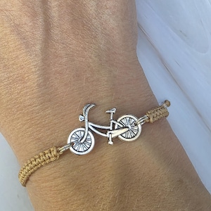 May include: A bracelet featuring a silver-toned metal bicycle charm centered on a woven golden-brown cord. The bicycle charm displays detailed wheels, a frame, and handlebars. The cord is secured with adjustable braided sections on both sides.
