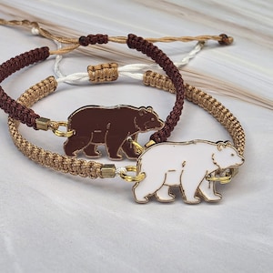 May include: Two adjustable cord bracelets featuring bear-shaped charms. One bracelet has a dark brown braided cord with a dark brown bear charm, while the other has a metallic gold braided cord with a white bear charm and a white pull cord.