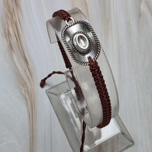 May include: A silver-toned bracelet featuring a cowboy hat design. The hat is detailed with a rope-like border and a central oval. The bracelet is strung on a dark brown woven cord, creating a rustic aesthetic.