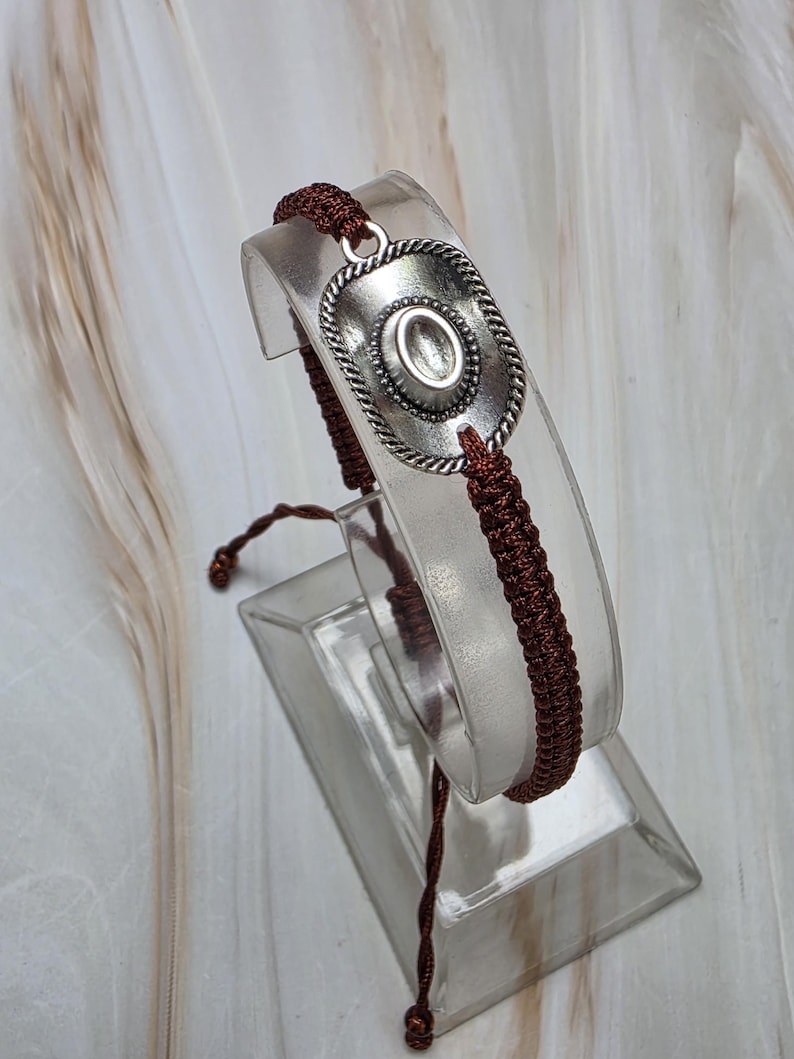 May include: A silver-toned bracelet featuring a cowboy hat design. The hat is detailed with a rope-like border and a central oval. The bracelet is strung on a dark brown woven cord, creating a rustic aesthetic.