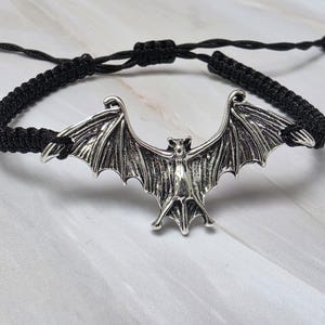 Puede incluir: Una pulsera de cordón trenzado negro con un dije de murciélago de metal en tono plateado. El murciélago se muestra con las alas extendidas, con texturas detalladas en el cuerpo y las alas. La pulsera cuenta con un cierre de nudo corredizo ajustable.
