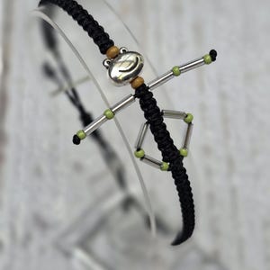 May include: A black macrame bracelet featuring a silver frog charm with wooden beads. The bracelet has silver rods and green beads forming arms and legs. The frog charm is the focal point of this unique, handmade accessory.