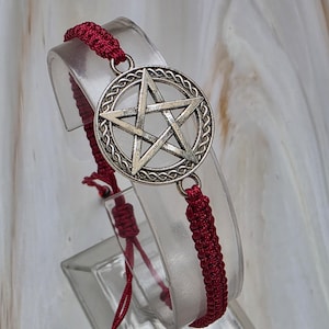 May include: A red macrame bracelet featuring a silver-colored pentagram charm. The charm is a five-pointed star within a circle with a braided border. The bracelet is displayed on a clear, curved stand.
