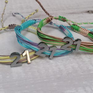 Puede incluir: Tres pulseras de amistad coloridas con dijes de metal plateado que dicen "21". Las pulseras están hechas de hilos tejidos en tonos de amarillo, verde, azul y marrón.