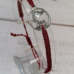 May include: A red braided bracelet with a silver charm featuring a mushroom and a small owl.