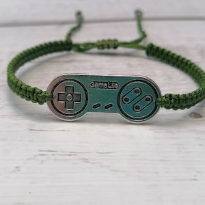 May include: A green cord bracelet with a silver charm shaped like a video game controller. The charm has the text "Game Life" on it.