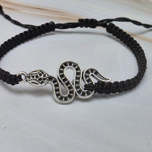 May include: A black cord bracelet featuring a silver-toned metal snake charm. The bracelet is constructed with a braided cord design and includes adjustable sliding knots at the ends to secure the fit around the wrist for comfortable wear.