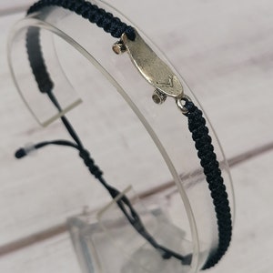 Puede incluir: Una pulsera trenzada negra con un dije de plata en forma de patineta. El dije tiene un peque&ntilde;o detalle en la parte superior de la tabla.