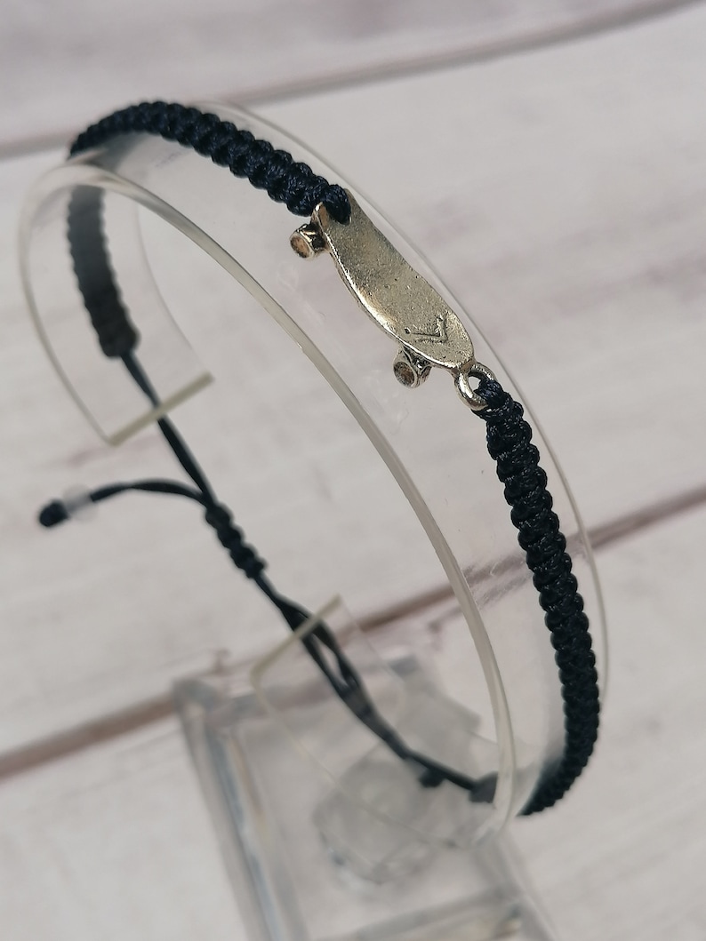 Puede incluir: Una pulsera trenzada negra con un dije de plata en forma de patineta. El dije tiene un peque&ntilde;o detalle en la parte superior de la tabla.