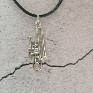 Puede incluir: Un colgante de trombón plateado cuelga de un collar de cordón negro. El dije de trombón es detallado, con correderas y campana visibles. El fondo es una superficie texturizada de color gris claro.