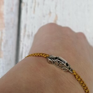 Puede incluir: Pulsera de macram&eacute; dorada con un charm de coche plateado. La pulsera se lleva en la mu&ntilde;eca, mostrando el intrincado anudado y el colgante de coche detallado. El fondo es una superficie de madera clara.