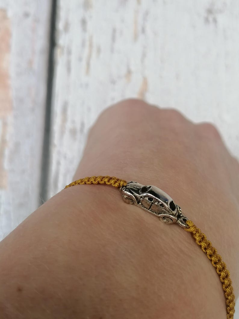 Puede incluir: Pulsera de macram&eacute; dorada con un charm de coche plateado. La pulsera se lleva en la mu&ntilde;eca, mostrando el intrincado anudado y el colgante de coche detallado. El fondo es una superficie de madera clara.