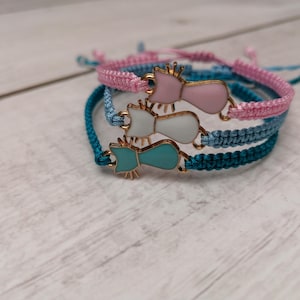 Pode incluir: Três pulseiras de amizade trançadas com pingentes de gato. As pulseiras são turquesa, azul e rosa.