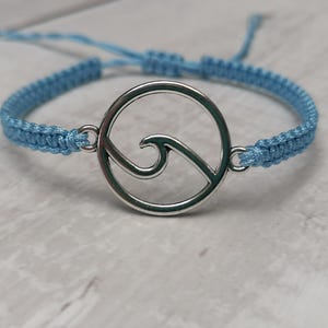 Puede incluir: Una pulsera trenzada azul claro con un dije plateado con diseño de ola.