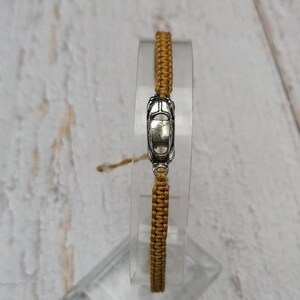 Puede incluir: Una pulsera tejida de color dorado con un dije de coche plateado. La pulsera se exhibe sobre un soporte transparente y rectangular, sobre un fondo claro de vetas de madera. El dije es una miniatura detallada de un coche cl&aacute;sico.