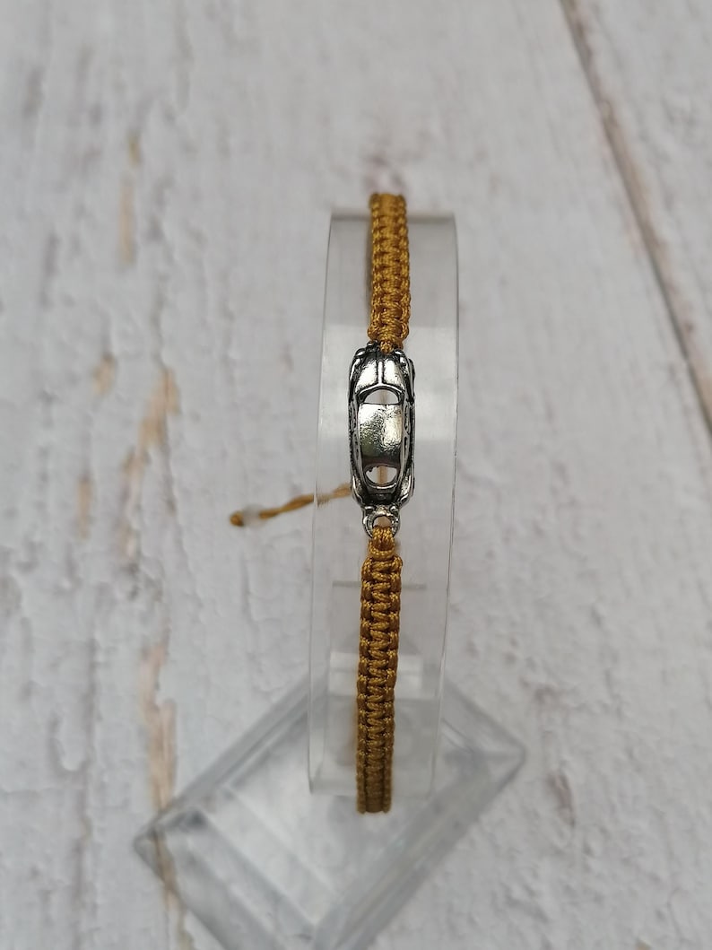 Puede incluir: Una pulsera tejida de color dorado con un dije de coche plateado. La pulsera se exhibe sobre un soporte transparente y rectangular, sobre un fondo claro de vetas de madera. El dije es una miniatura detallada de un coche cl&aacute;sico.
