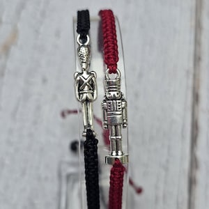 May include: Two adjustable bracelets with silver-colored charms. One bracelet is black, the other is red. The charms are shaped like a soldier and a nutcracker. The bracelets are displayed on a clear stand against a light wood background.